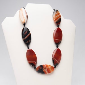 May include: A necklace made of oval-shaped agate beads in shades of red, orange, and black. The beads are strung on black cord.