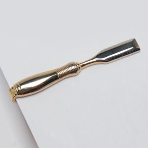 May include: A gold-toned metal tie clip shaped like a woodworking chisel. The accessory features a smooth, reflective handle with a textured grip section and a flat, rectangular blade end, designed to secure a necktie with a functional clasp.