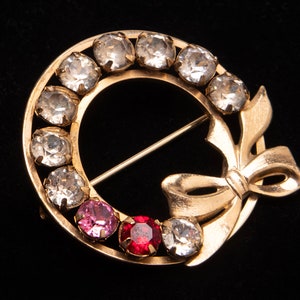 May include: A gold-toned brooch in the shape of a wreath with a bow. The wreath is adorned with clear rhinestones and the bow has red and pink rhinestones.