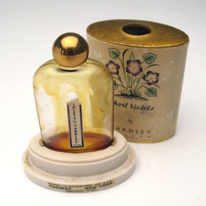 May include: A vintage glass bottle of Yardley's April Violets perfume. The bottle is clear and has a gold cap. The bottle is sitting on a white pedestal. The bottle is in its original box, which is white with a gold top and a floral design. The box says "April Violets by Yardley".