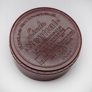 May include: A circular, dark brown container with the text "Linda Neutral" in a stylized font. The container has a screw-on lid and includes additional text in German. The container holds 150 grams of product.