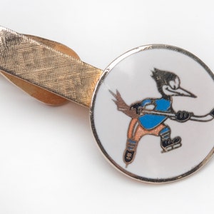 Peut inclure: Un fermoir de cravate en or avec un cercle en émail blanc représentant un pic-bois de dessin animé portant un maillot bleu et un short orange, tenant un bâton de hockey. Le pic-bois patine sur la glace.