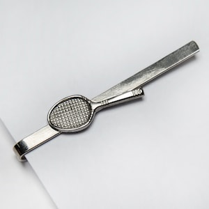 May include: Silver-toned tie clip featuring a tennis racket design. The clip has a rectangular bar with a curved end and a detailed racket charm. The racket's strings are visible, and the clip is set against a white background.
