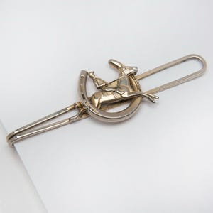 May include: Silver-toned tie clip featuring a horse and rider jumping over a horseshoe. The clip has a rectangular bar and a decorative equestrian design. The horse and rider are in mid-air, adding a dynamic element to the accessory.