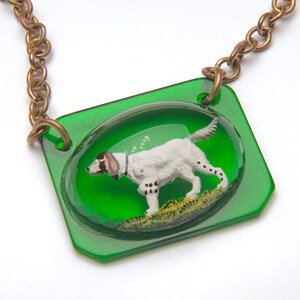 May include: A green pendant necklace with a brass chain features a white dog with black spots inside a clear oval. The dog is depicted running through green grass.