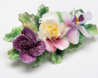 Peut inclure: Une broche en porcelaine en forme de fleurs avec un arrangement de fleurs violettes, roses et jaunes. Les fleurs sont entourées de feuilles vertes.