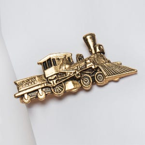 May include: A gold-colored train pin with the text "C&NW Pioneer" on the side. The pin features a detailed design of a classic steam engine, including the smokestack, cab, and wheels. The pin is likely made of metal.
