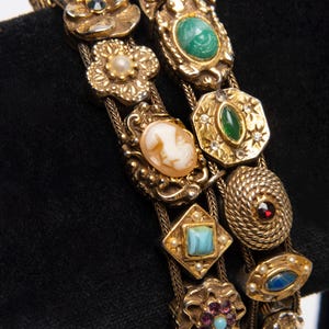 May include: Close-up of two gold-toned bracelets with ornate, vintage-style charms. The charms include floral designs, cameos, and various gemstones in shades of green, blue, and red. The bracelets are displayed against a black background.