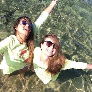 Peut inclure: Deux femmes portant des lunettes de soleil et des chemises vert clair sourient et se tiennent dans l'eau peu profonde. L'eau est claire et on peut voir le fond.