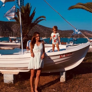 Peut inclure: Une femme en robe blanche se tient sur un bateau blanc avec un jeune enfant en maillot de bain rouge et blanc. Le bateau est amarré à un quai avec un ciel bleu et des palmiers en arrière-plan. Le bateau porte l'inscription "ΑΓΑΠΗ" sur le côté.