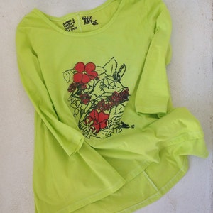 Peut inclure: Un t-shirt à manches longues vert lime avec un imprimé floral en rouge et noir. Le t-shirt a un col rond et un ourlet arrière plus long. Le texte "Designed & Printed by Christine La Smyth" et "Blue Sky Arts" est imprimé sur le t-shirt.