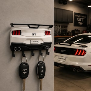May include: A white and black Mustang GT key holder mounted on a gray wall, featuring a spoiler, taillights, and exhaust pipes. Two car keys hang from the key holder. A white Mustang GT car is visible in the background.