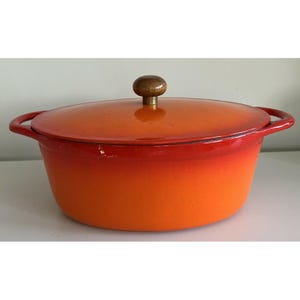 May include: An oval enameled cast iron Dutch oven featuring a vibrant gradient finish that transitions from a deep red at the rim to a bright orange at the base. It includes two side handles and a matching lid topped with a round wooden knob.