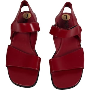 May include: A pair of red slingback sandals featuring a wide front strap and an adjustable ankle strap. The footbed is marked with the text BCBG and a circular sticker displaying the number 7. The sandals have a smooth finish and black soles.