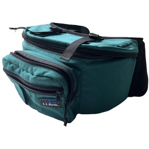 May include: A dark teal waist pack featuring black zippers, black adjustable straps, and a black fabric back panel. The front pocket displays a rectangular patch with the text L.L.Bean in white letters and a small graphic of a mountain range above it.