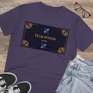 Könnte beinhalten: Ein lila T-Shirt mit einem marineblauen Rechteck mit dem Text "TEAM PFIZER" und "Est. 2021". Das Rechteck hat goldene Zierecken und zwei blau-weiße Pflaster. Ein Paar schwarze Sneaker, eine Brille und Jeans sind ebenfalls zu sehen.
