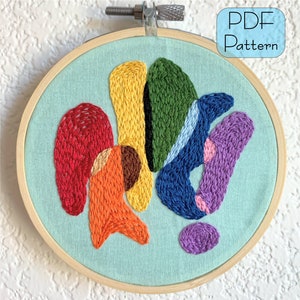May include: A colourful abstract embroidery pattern in a wooden embroidery hoop. The design features a variety of shapes and colours, including red, orange, yellow, green, blue, and purple.