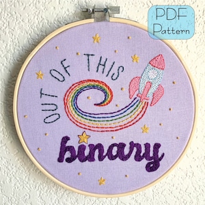 Peut inclure: Art de broderie sur un cercle avec un fond violet, un arc-en-ciel, une fusée et le texte "Out of this binary".