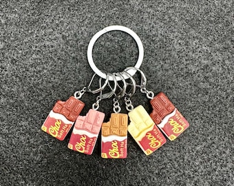 May include: Five chocolate bar keychains with a silver key ring. The keychains are different colors and have the text "Choc Soft Ha" on them.