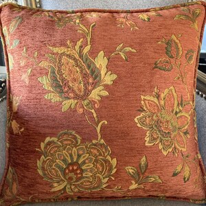 May include: A rust-colored throw pillow with a floral pattern in shades of green, yellow, and brown. The pillow has a textured fabric and a subtle sheen.