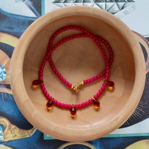 Puede incluir: Un collar con pequeñas cuentas de color rosa vibrante ensartadas con un cierre de tono dorado. El collar está adornado con cinco dijes colgantes de color ámbar en forma de lágrima, espaciados uniformemente a lo largo de la cadena.