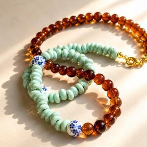 Könnte beinhalten: Zwei Armbänder mit Perlen. Ein Armband besteht aus bernsteinfarbenen Perlen, das andere aus hellgrünen Perlen. Das hellgrüne Armband hat weiße und blaue Perlen mit Blumenmuster. Das bernsteinfarbene Armband hat einen goldenen Verschluss.