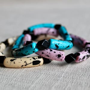 May include: Three bracelets made of cylindrical beads in turquoise, light pink, and cream, each featuring a black speckled pattern. The bracelets are accented with small, solid black square beads interspersed between the longer cylindrical pieces.