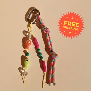 Puede incluir: Un llavero con un mosquetón de metal plateado unido a un cordón trenzado multicolor y dos cuerdas amarillas. Una cuerda tiene cuentas transparentes, naranjas con forma de pez, verdes y verde lima con forma de pez. La otra tiene cuentas rosas y verdes.