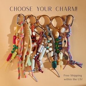 Puede incluir: Una colección de seis llaveros con anillos y mosquetones de metal dorado, adornados con cordones de nailon de colores, cuentas de vidrio y cerámica variadas, y pequeños dijes en forma de pez. El texto CHOOSE YOUR CHARM! es visible arriba.