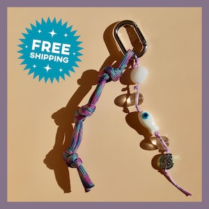May include: A keychain featuring a silver-toned metal carabiner attached to a multicolored paracord with three knots and a separate strand of beads including a white fish-shaped evil eye charm, clear glass spheres, and a small metallic disc.