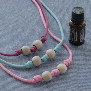 May include: Three knotted cords with wooden beads, each in a different color: pink, blue, and burgundy. A small bottle of essential oil blend is in the background.