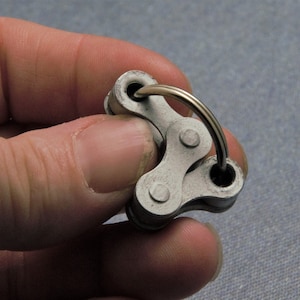 May include: A silver metal keychain with a chain link design. The keychain is held in a hand.