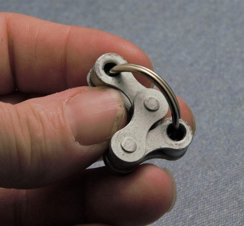 May include: A silver metal keychain with a chain link design. The keychain is held in a hand.