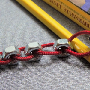 May include: A red cord threaded through a series of silver metal nuts, creating a chain-like design. The cord is wrapped around a yellow pencil.