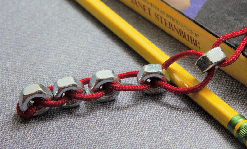May include: A red cord threaded through a series of silver metal nuts, creating a chain-like design. The cord is wrapped around a yellow pencil.