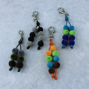 May include: Four keychains with different colored pom-pom beads. The keychains are made with a silver metal clip and a brown cord. The beads are gray, black, blue, green, orange, and brown.