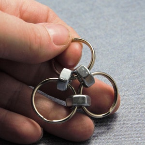 Puede incluir: Un llavero de metal plateado con tres anillos entrelazados y tres tuercas plateadas. El llavero est&aacute; sostenido en una mano.