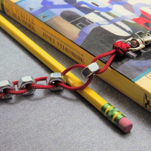 May include: A red and silver keychain with a silver carabiner clip and a chain of silver nuts. The keychain is lying on top of a yellow book with the title "The Witch in Her Work" by Janet Sternburg. A yellow pencil with green accents is lying next to the keychain.