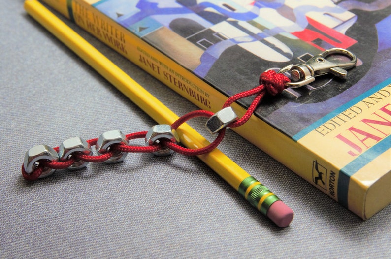 May include: A red and silver keychain with a silver carabiner clip and a chain of silver nuts. The keychain is lying on top of a yellow book with the title "The Witch in Her Work" by Janet Sternburg. A yellow pencil with green accents is lying next to the keychain.