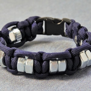 May include: A dark blue paracord bracelet with silver metal nuts woven into the design. The bracelet has a black plastic buckle closure.