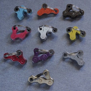 May include: Nine different colored bike chain keychains. The keychains are made of metal and have a ring at the top. The colors are blue, orange, black, pink, white, yellow, purple, silver, and gold.