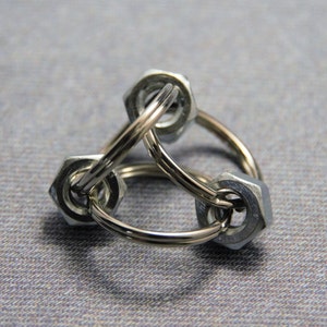 Puede incluir: Un llavero de metal plateado hecho de tres tuercas conectadas. Las tuercas est&aacute;n unidas por un anillo de metal plateado.