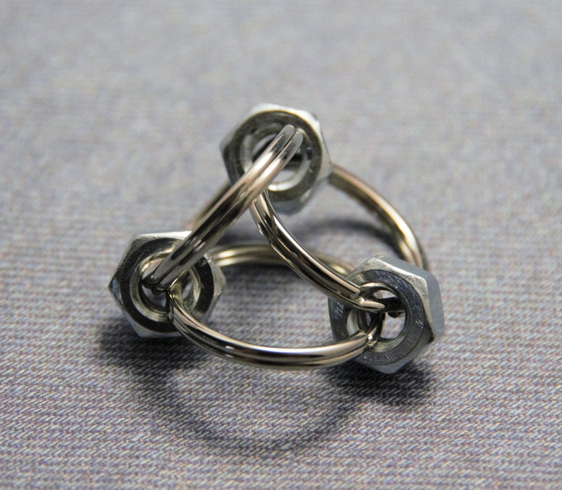 Puede incluir: Un llavero de metal plateado hecho de tres tuercas conectadas. Las tuercas est&aacute;n unidas por un anillo de metal plateado.