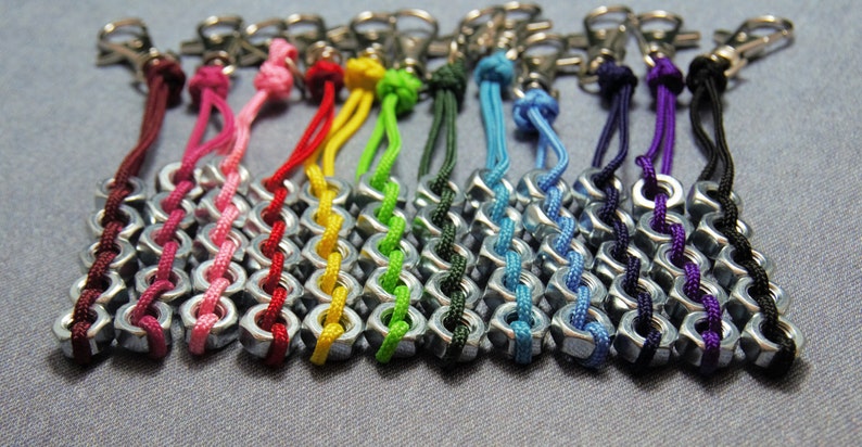 May include: A collection of colorful keychains made with nuts and string. The keychains are in various colors, including red, pink, yellow, green, blue, purple, and black.