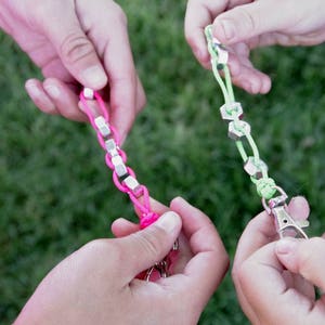 May include: Three hands holding colorful string bracelets with metal accents. The bracelets are made with pink, green, and silver colored string and metal.