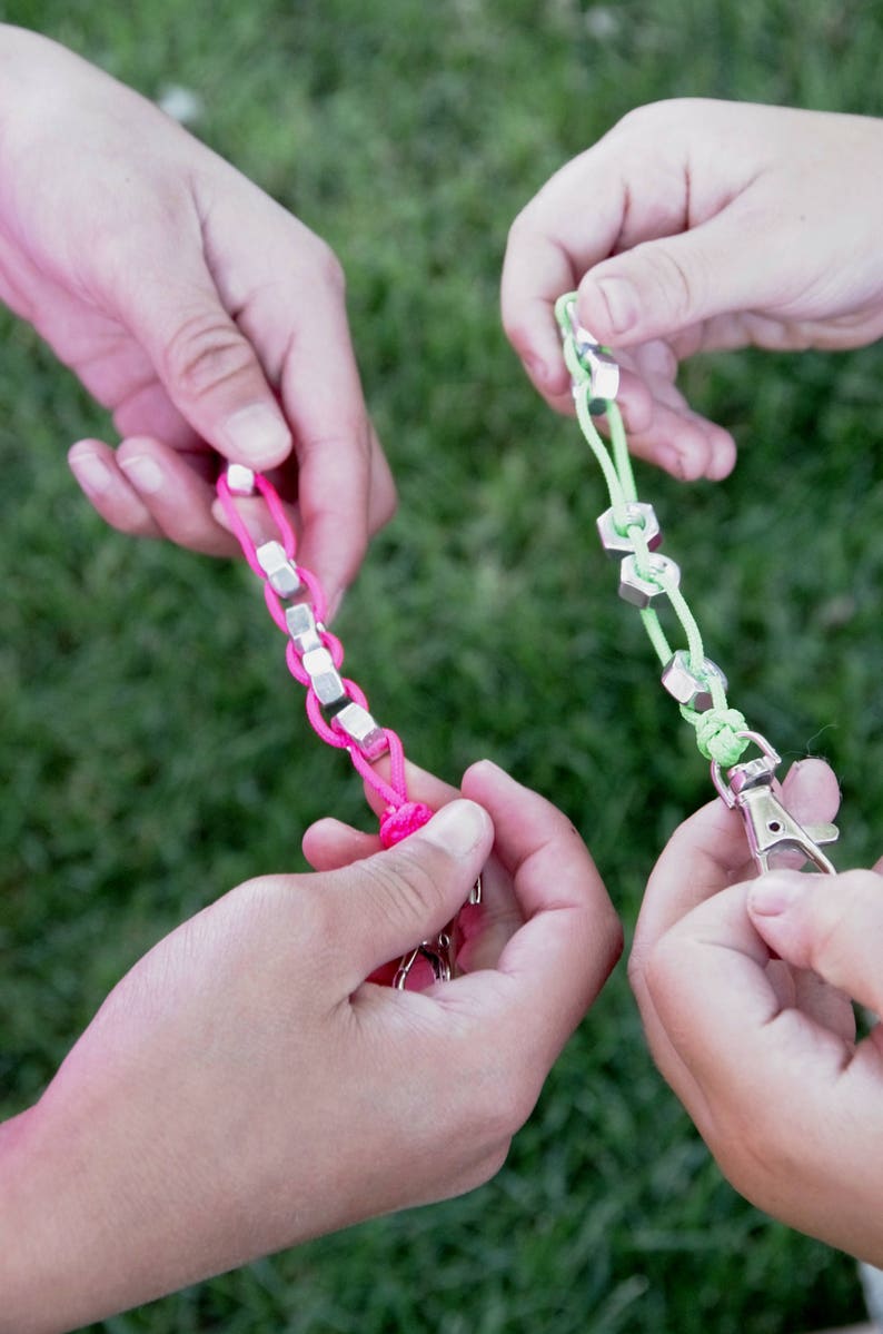 May include: Three hands holding colorful string bracelets with metal accents. The bracelets are made with pink, green, and silver colored string and metal.