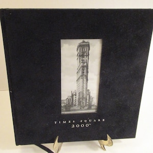 May include: Black photo album with a framed black and white image of a tall building. The text "TIMES SQUARE 2000" is printed in silver below the image. The album is displayed on a gold-colored stand.