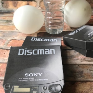 May include: A black Sony Discman CD player with the text "SONY 10 PLAYBACK MODES CD COMPACT PLAYER D-121 MEGA BASS" printed on the front. The player is sitting on a brick surface.