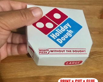 Puede incluir: Una caja hexagonal blanca con el logotipo de Domino's Pizza en rojo y azul, con el texto "Holiday Dough". La caja también incluye las frases "What's Pizza Without The Dough?" y "LARGE."