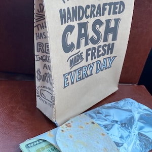 May include: A brown paper bag with the text "OUR HANDCRAFTED CASH MADE FRESH EVERY DAY" in black. A wrapped burrito and a twenty-dollar bill are in front of the bag.